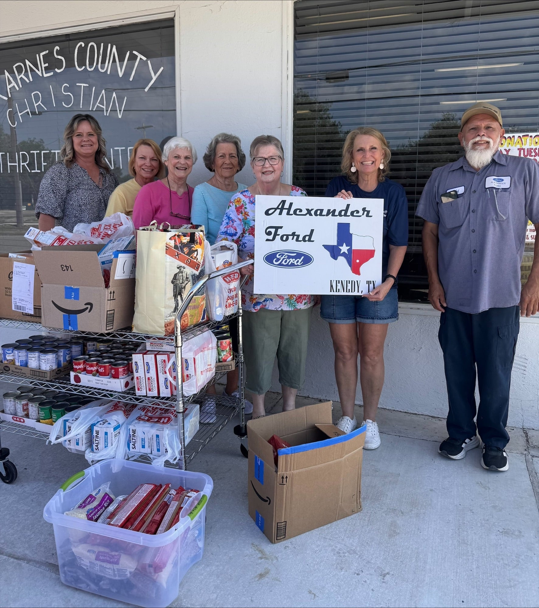 Group of seven smiling people next to a cart of donated food, holding an 'Alexander Ford Kenedy, TX' sign.